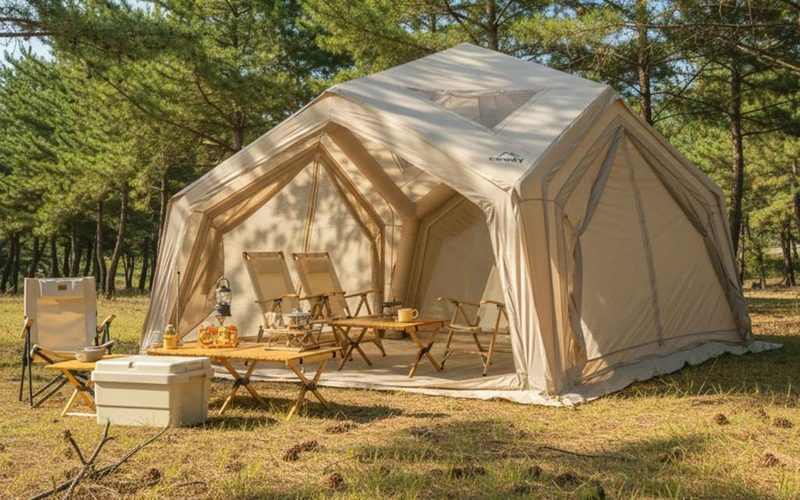 Inflatable camping tent with folding chairs and a table among pine trees